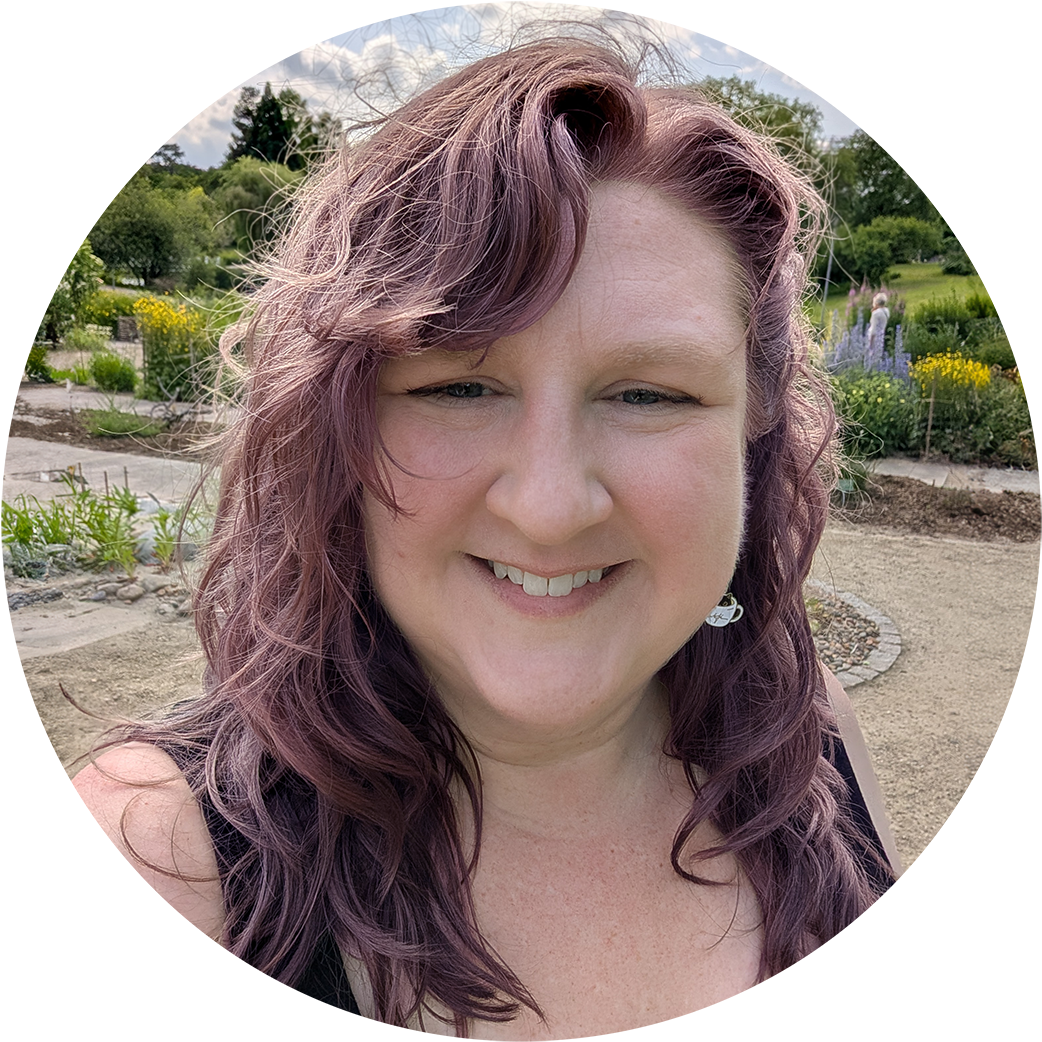 A selfie of Angela. A white woman with purple wavy hair in a wooded park.